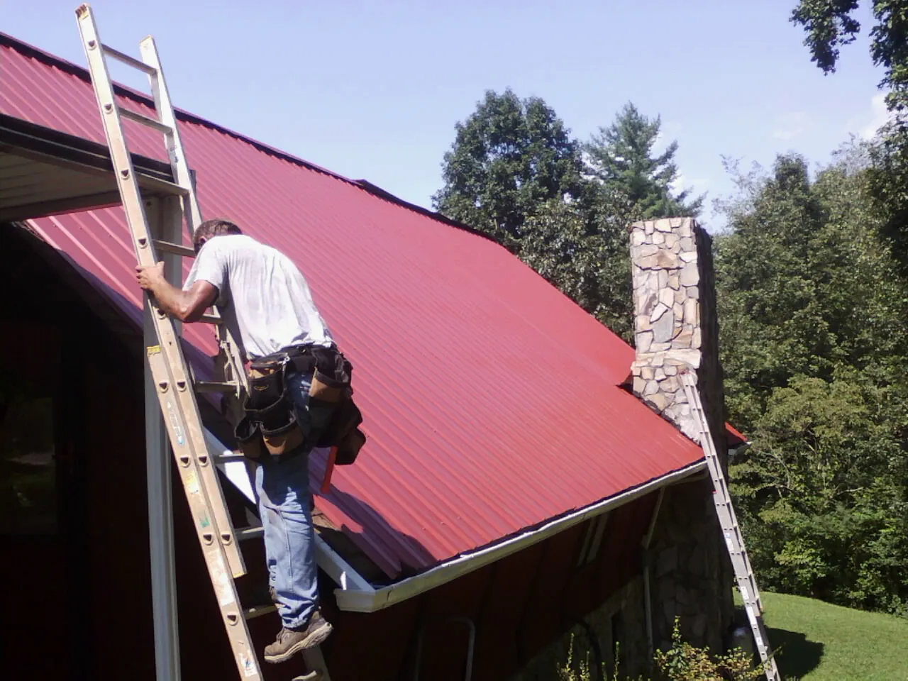 Roofer on a ladder beside a red metal roof and stone chimney — Soffit & Fascia Repair by Roofing Brothers in Gray